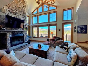 A spacious living room with a stone fireplace, large windows offering mountain views, a cozy seating area, and a chandelier. - MM1 Luxury Home, Walk to Mount Washington Hotel! AC, Heated Floor, Stunning Views Pool Gym! (Bretton Woods)