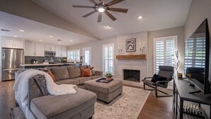 A spacious living room with a gray sofa, a coffee table, and a fireplace. There is a kitchen in the background with modern appliances and a dining area. - Central Chandler (Chandler)