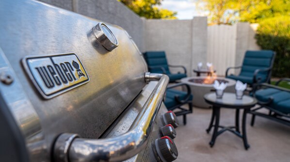 Un barbecue Weber en acier inoxydable, avec le logo de la marque bien visible, un foyer et un espace extérieur aménagé avec des chaises et une table.