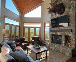 Un amplio salón con sofás de cuero, chimenea de piedra y ventanas grandes con vistas al exterior.