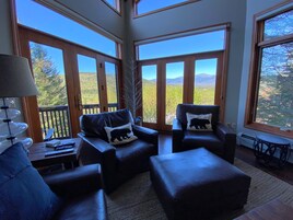 Una habitación acogedora con ventanas amplias, sofás de cuero con almohadones de temática de osos, una mesita con lámpara y vistas a un paisaje montañoso.