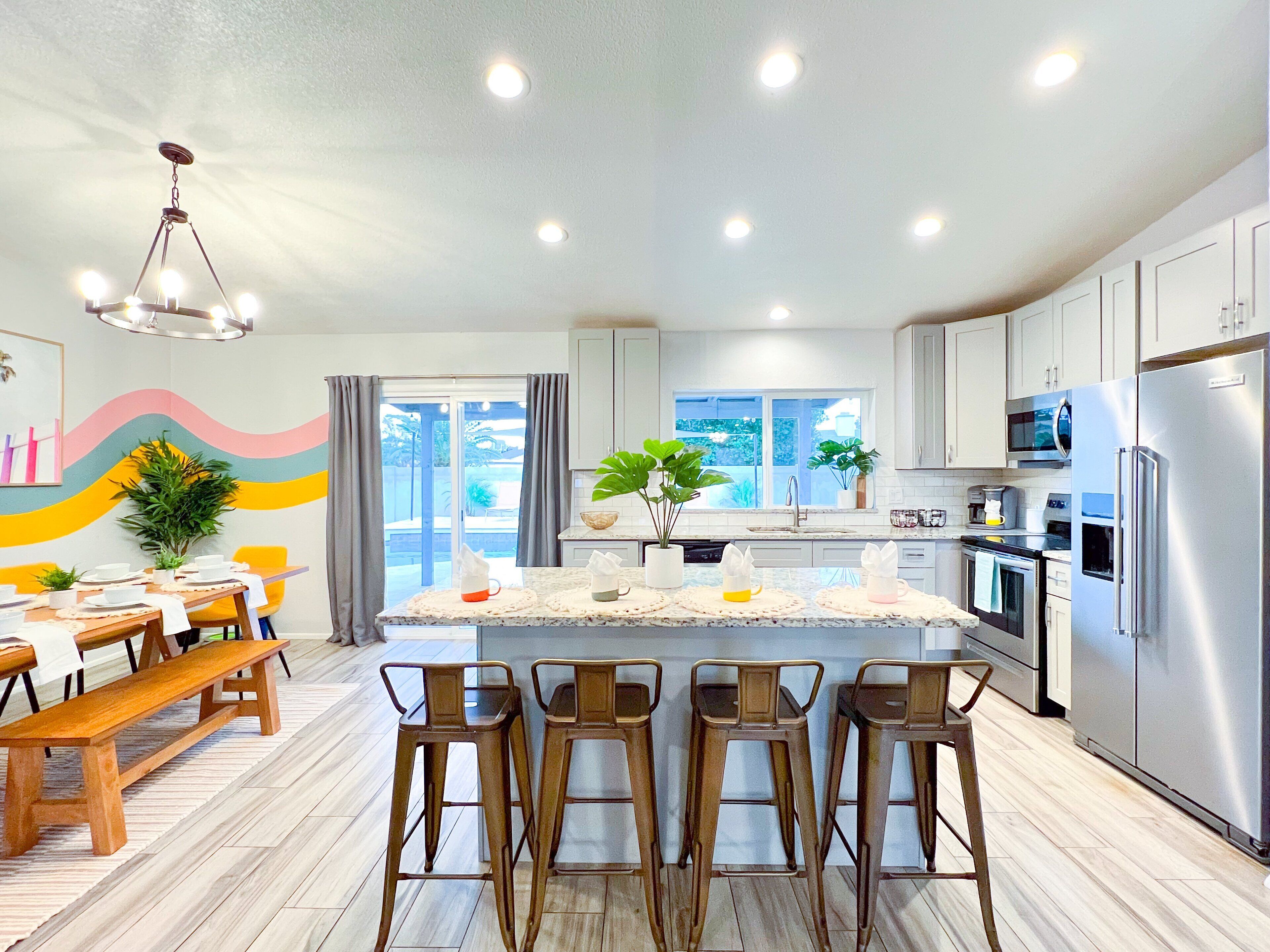 A modern kitchen with a central island, bar stools, a dining table with chairs, and a view to the outside.