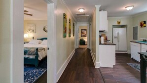 Un pasillo estrecho con una cama a la izquierda, una zona de cocina con refrigerador y fregadero a la derecha, y una puerta que da a otra habitación.