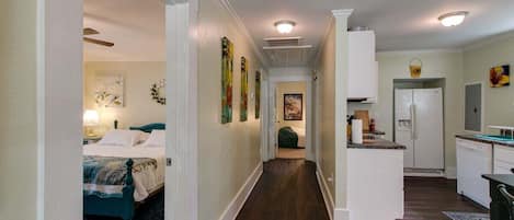 A narrow hallway with a bed on the left, a kitchen area with a refrigerator and sink on the right, and a door leading to another room.