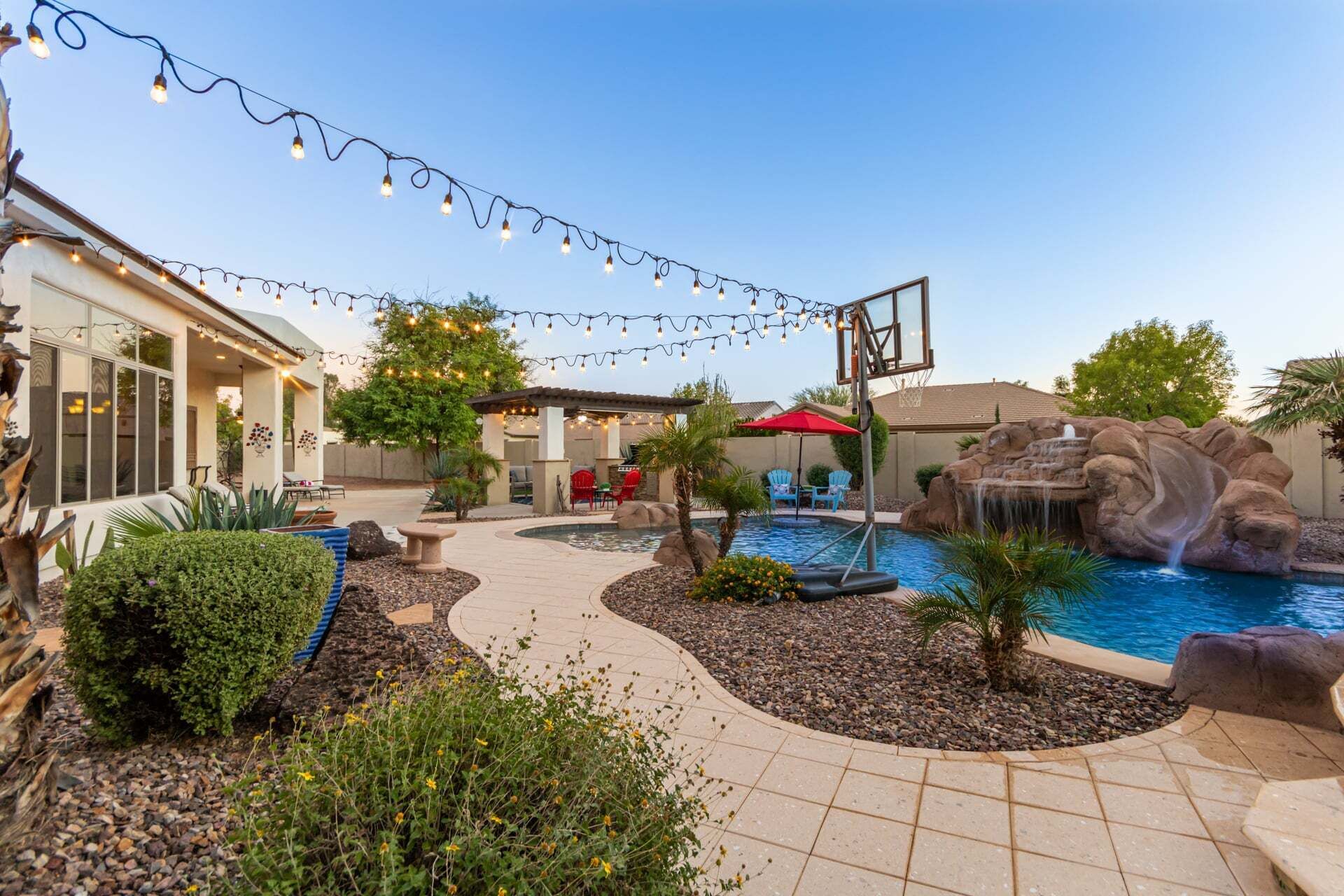 A backyard with a swimming pool, a basketball hoop, string lights, and a patio area.