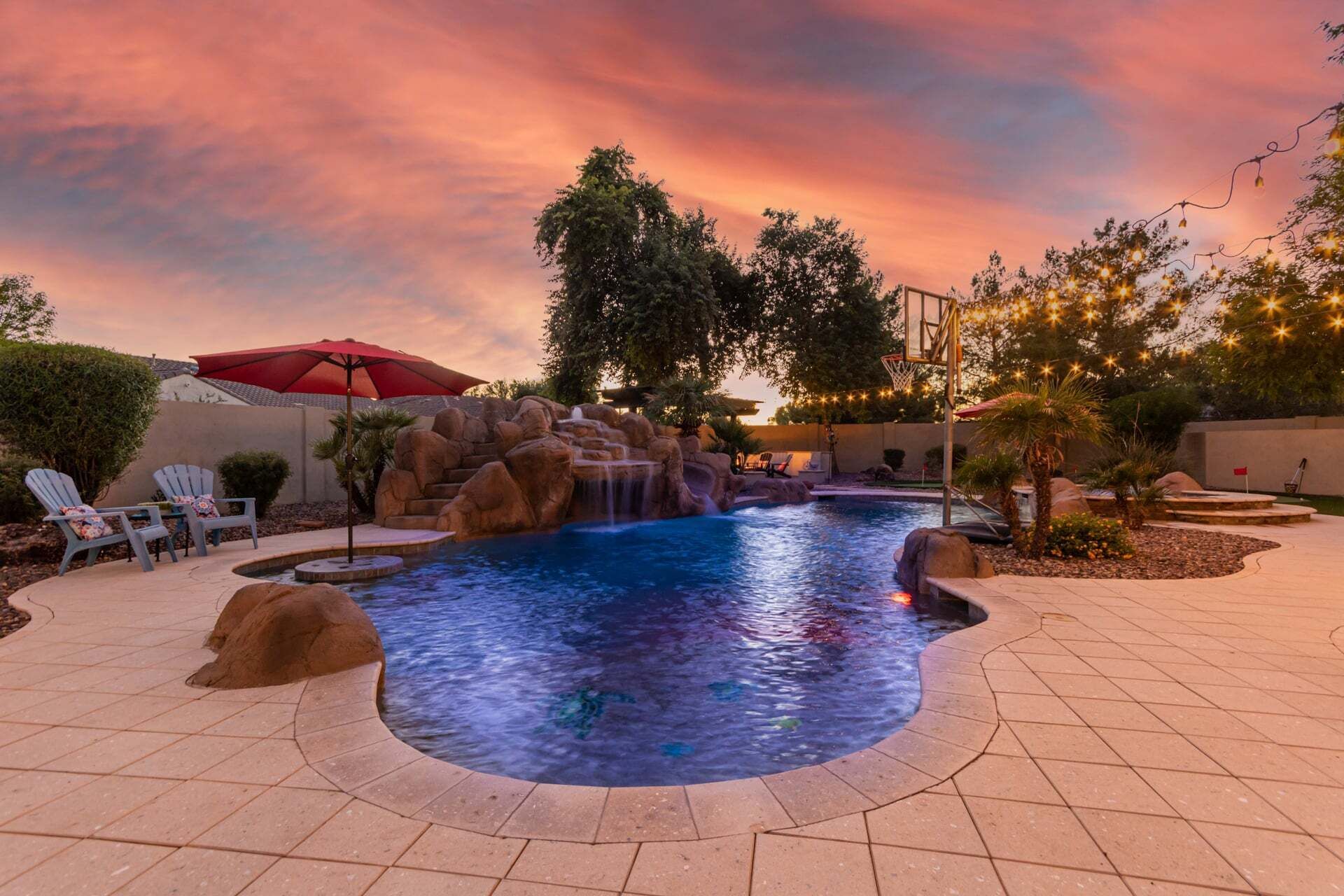 Piscina com cascata, rodeada por espreguiçadeiras e um guarda-sol vermelho.