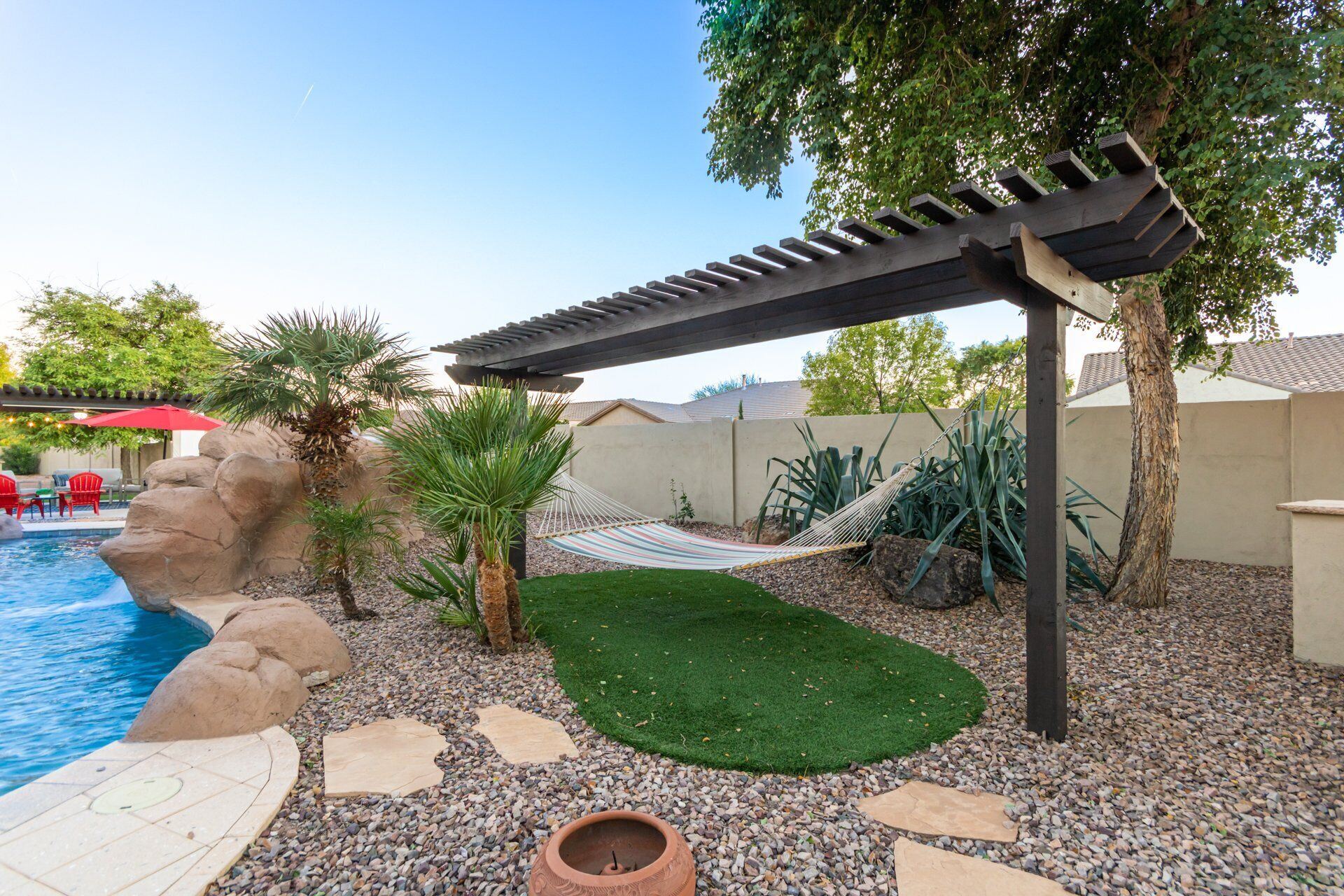 A backyard with a pool, palm trees, a hammock, and a pergola.