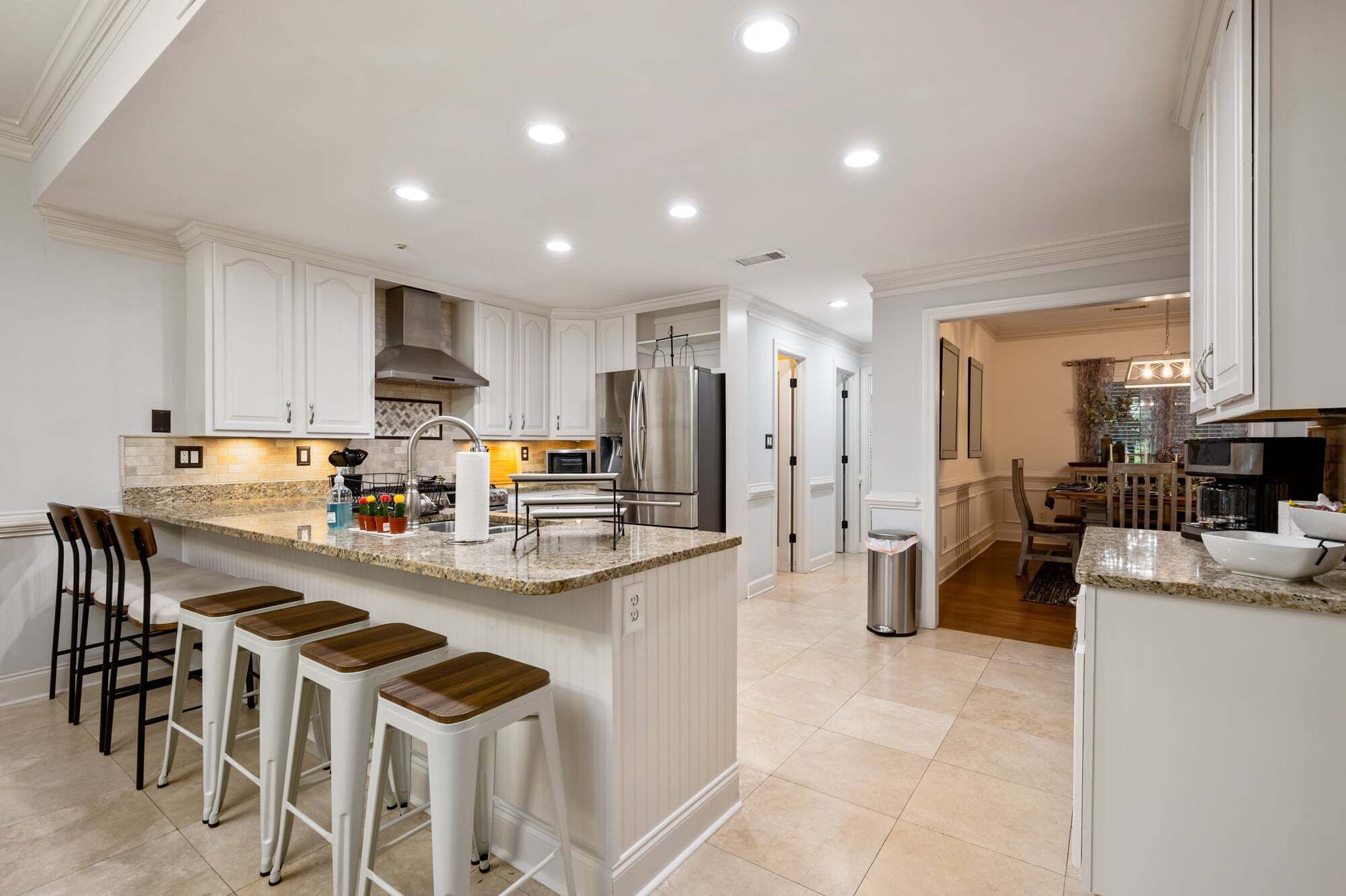 A modern kitchen with white cabinetry, a central island with stools, stainless steel appliances, and recessed ceiling lighting.
