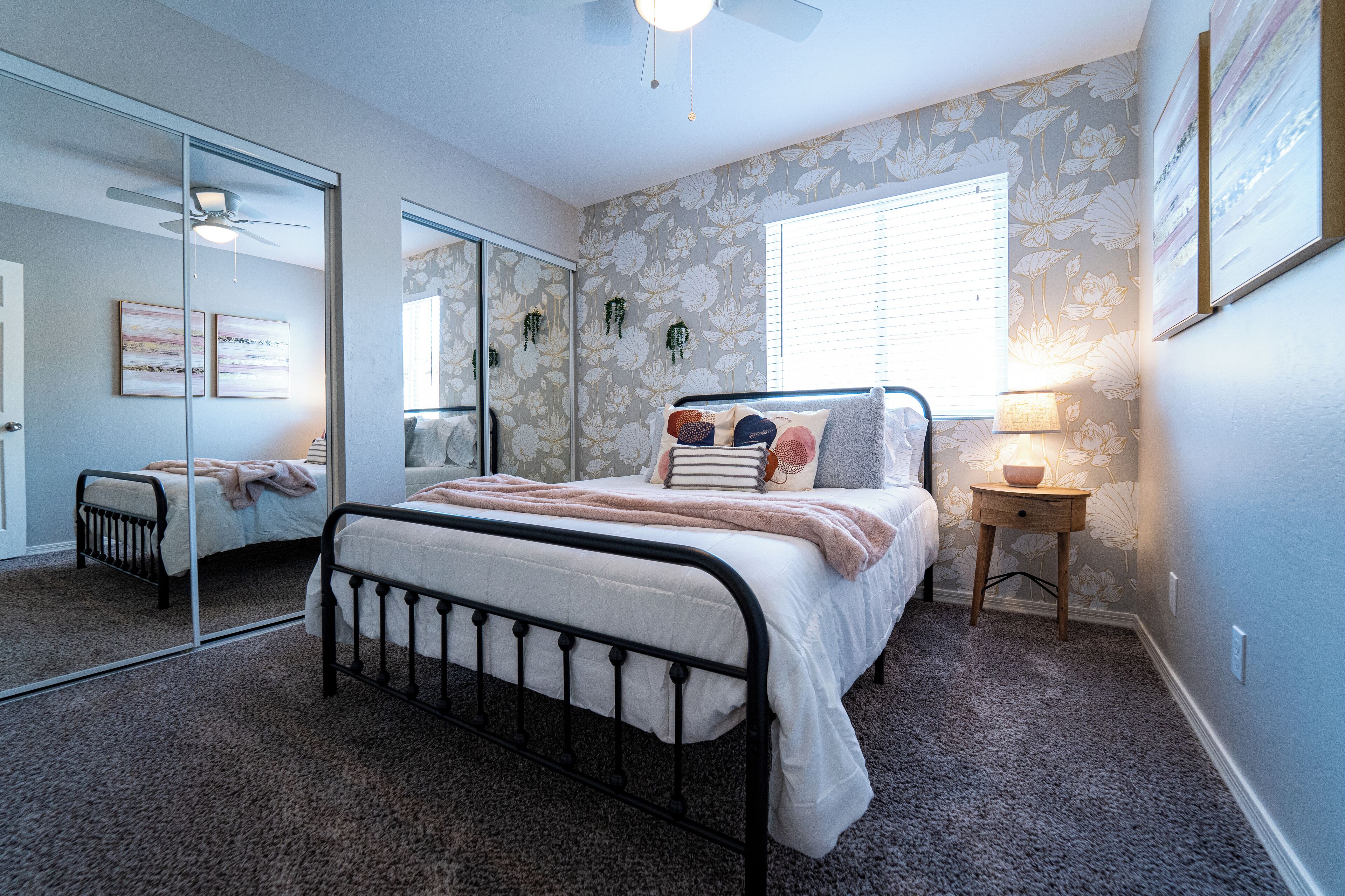 A bedroom with a bed, a bedside table with a lamp, a mirror, and a window with blinds.