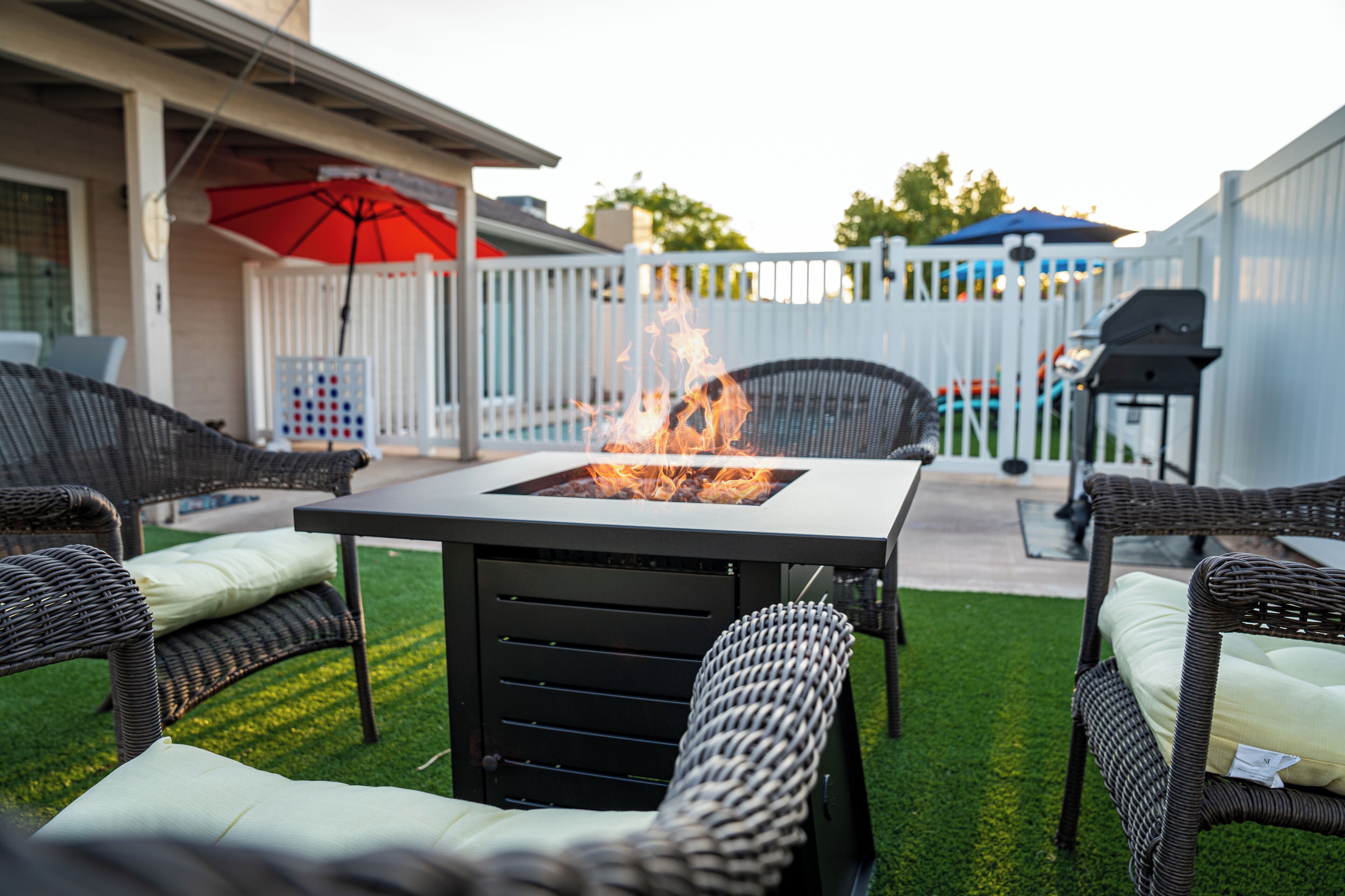Une terrasse extérieure avec des meubles en osier, une table carrée dotée d’un foyer, et une clôture blanche en arrière-plan.