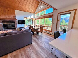 Amplia sala de estar con chimenea, televisor, mesa de comedor con sillas y ventanas grandes que ofrecen vistas al exterior.