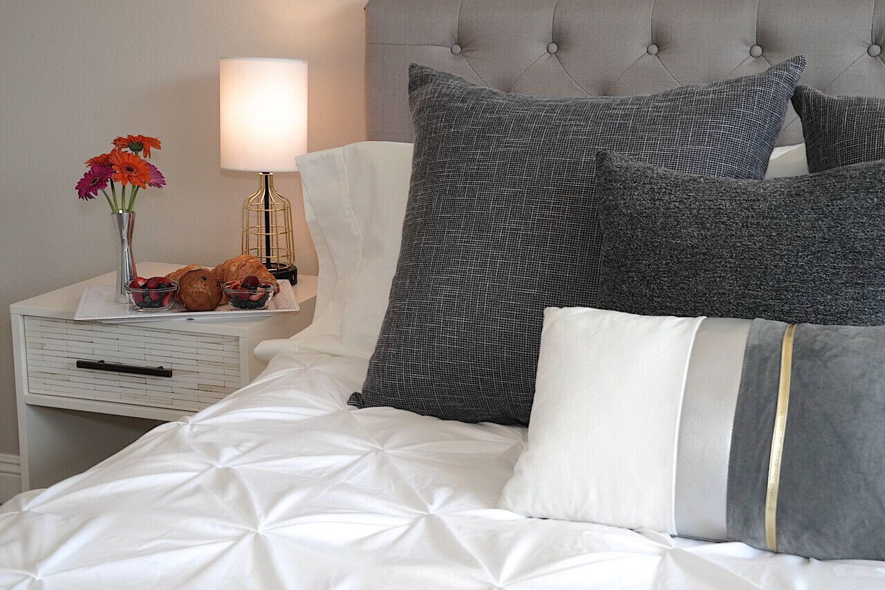 A neatly made bed with a white duvet, grey and white pillows, and a bedside table with a lamp, flowers, and croissants.