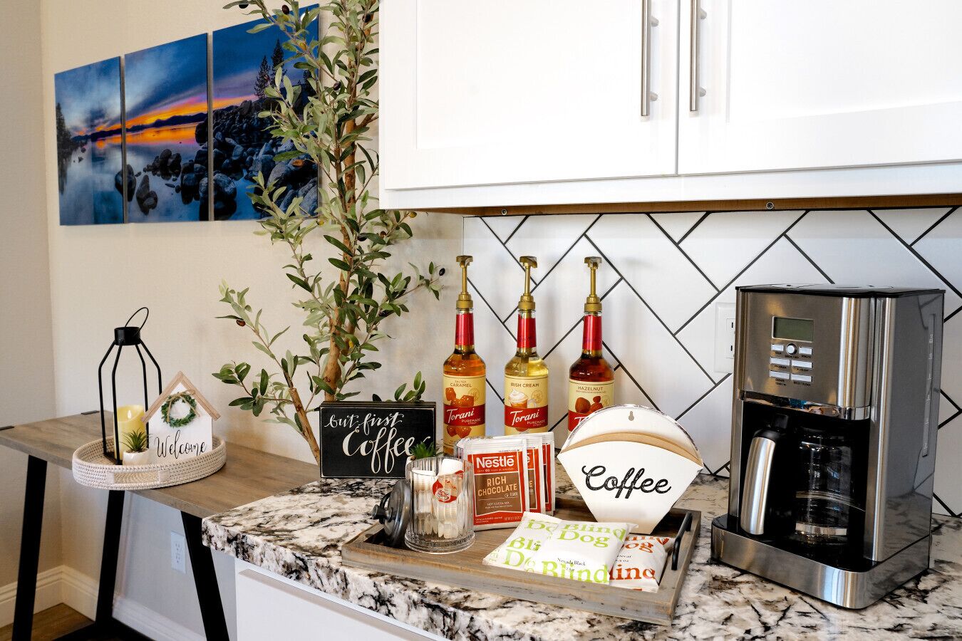 A kitchen counter with a coffee station, including a coffee maker, bottles of Torani syrups, Nestle chocolate bars, and a sign that says "but first Coffee".