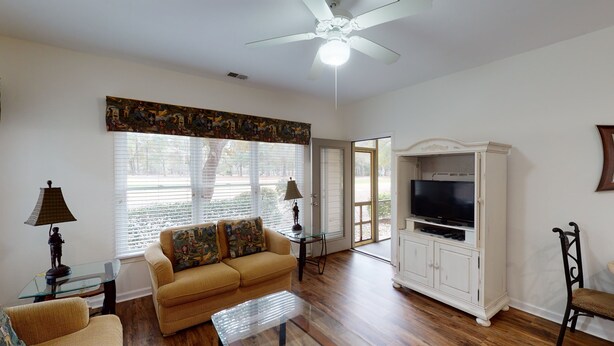 A living room with a sofa, a glass coffee table, a television on a cabinet, and a ceiling fan. - Club Villas Unit 23A - Lower Level (Sunset Beach)
