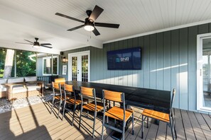 Ein überdachter Außenbereich mit Essbereich, einem langen Tisch, Stühlen, Deckenventilatoren und einem an der Wand montierten Fernseher.