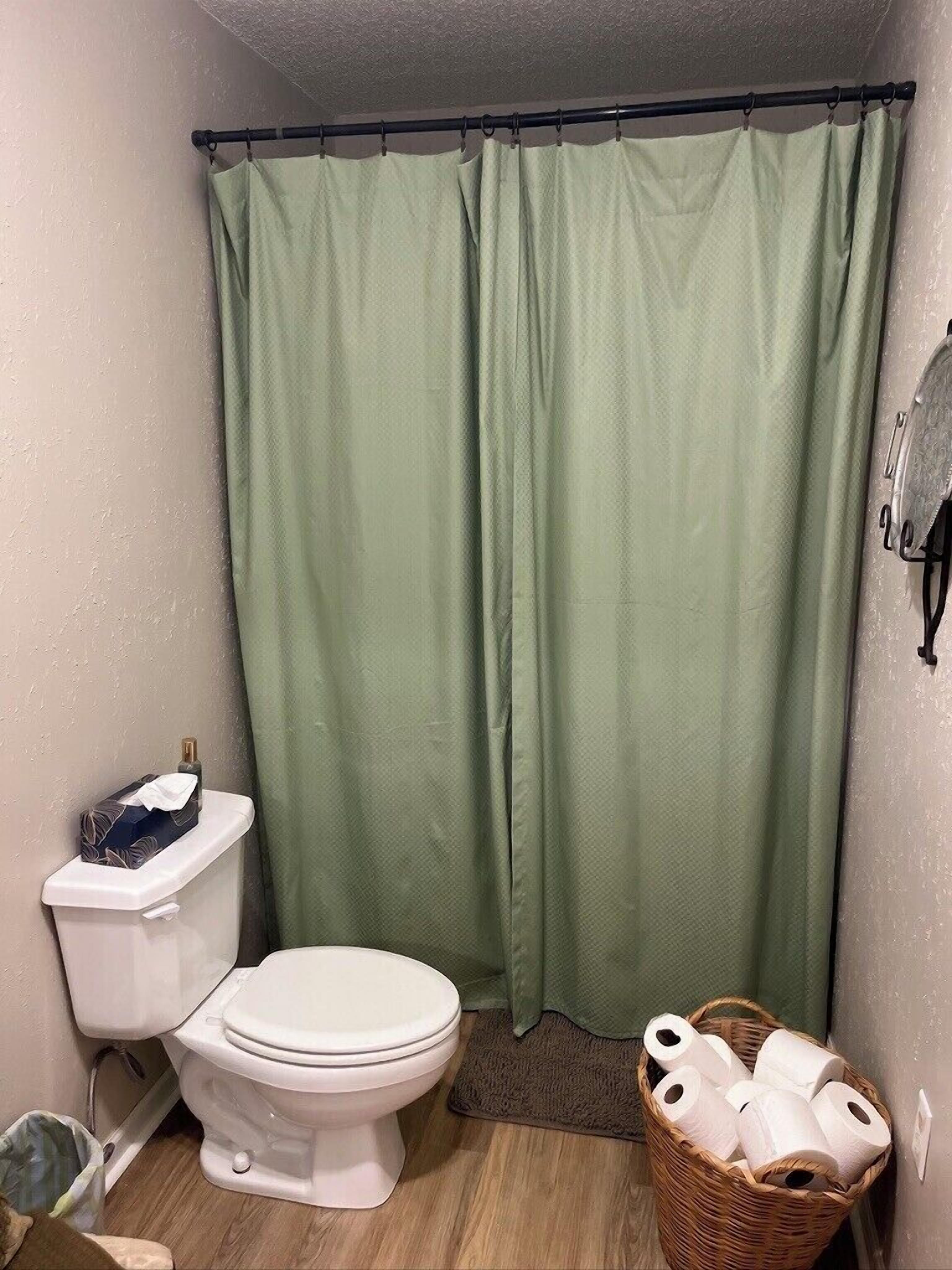 Casa de banho com sanita, cortina de chuveiro verde e uma cesta de papel higiénico.