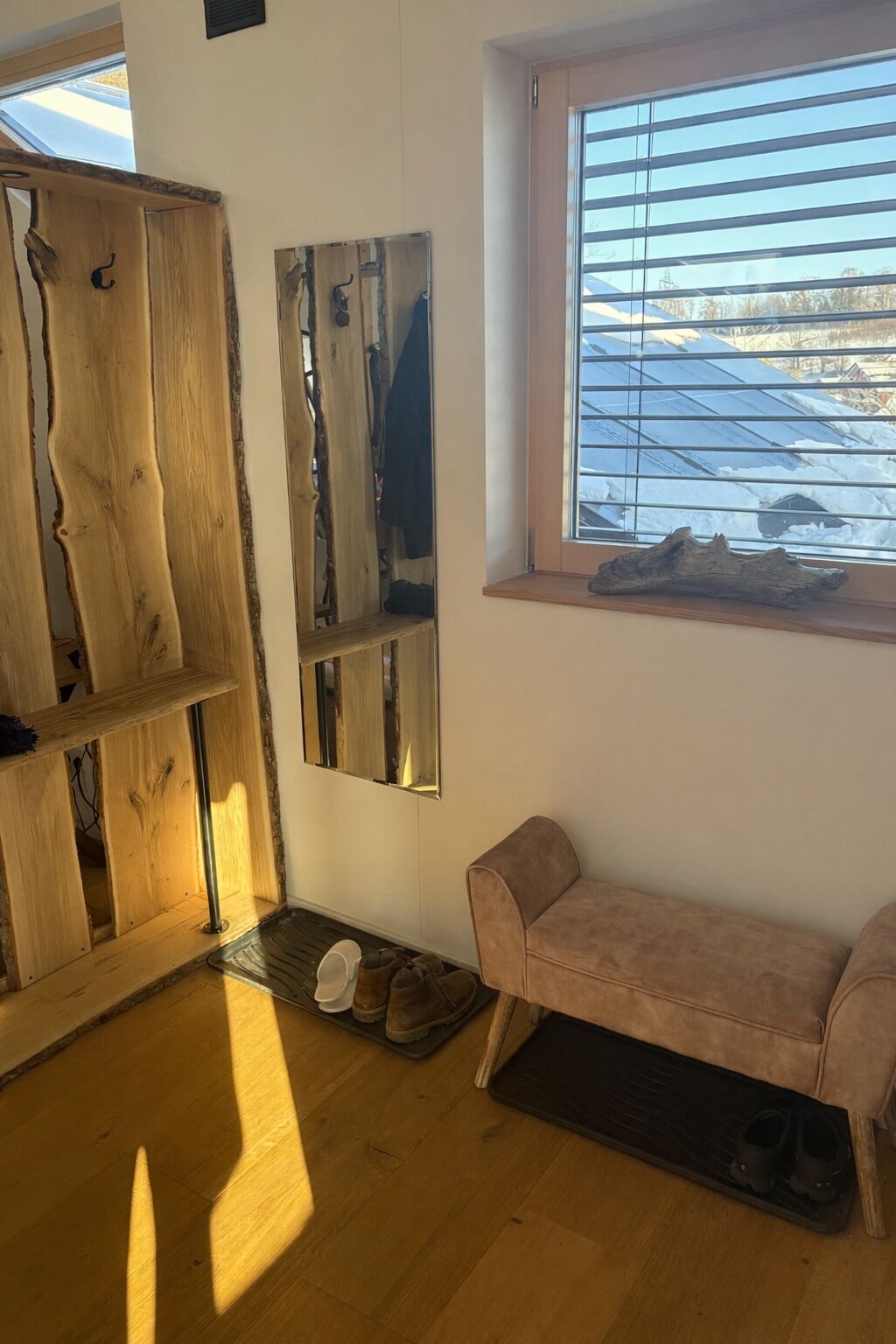 Una habitación con piso de madera, un banco, una ventana con persianas y un perchero.