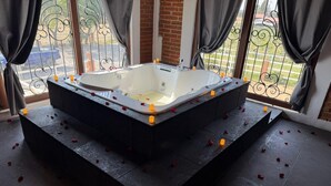 Ein Wellnessbereich mit einer großen Badewanne, Rosenblüten auf dem Boden verteilt und um die Fenster gezogenen Vorhängen.