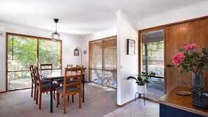 A dining area with a wooden table and chairs, large windows with blinds, and a potted plant on a stand.