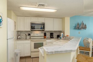 A compact kitchen with white cabinets, a microwave, an oven, and a coffee maker. There is a small trash bin and a wooden chair. - Panaramic oceanfront views abound lazy river pools kitchen washer dryer (Myrtle Beach)