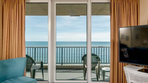 A room with a sliding glass door leading to a balcony with a view of the sea. There is a blue sofa, two green chairs, and a white TV stand. - Oceanfront resort with all amenities (Myrtle Beach)