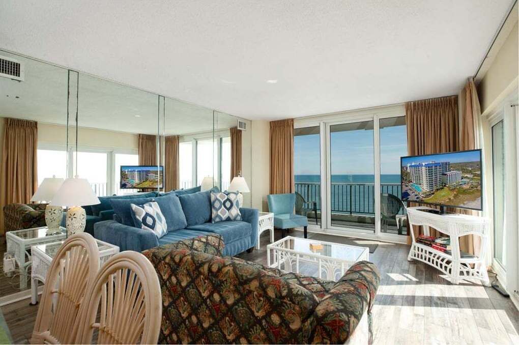 A spacious living room with a large sofa, a television, and a balcony with a view of the sea.