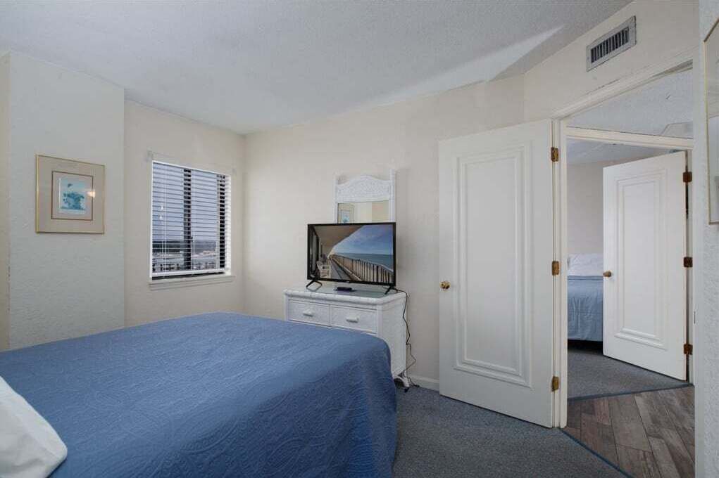 Ein Schlafzimmer mit Bett, Kommode, Fernseher und einer Tür zu einem anderen Raum.