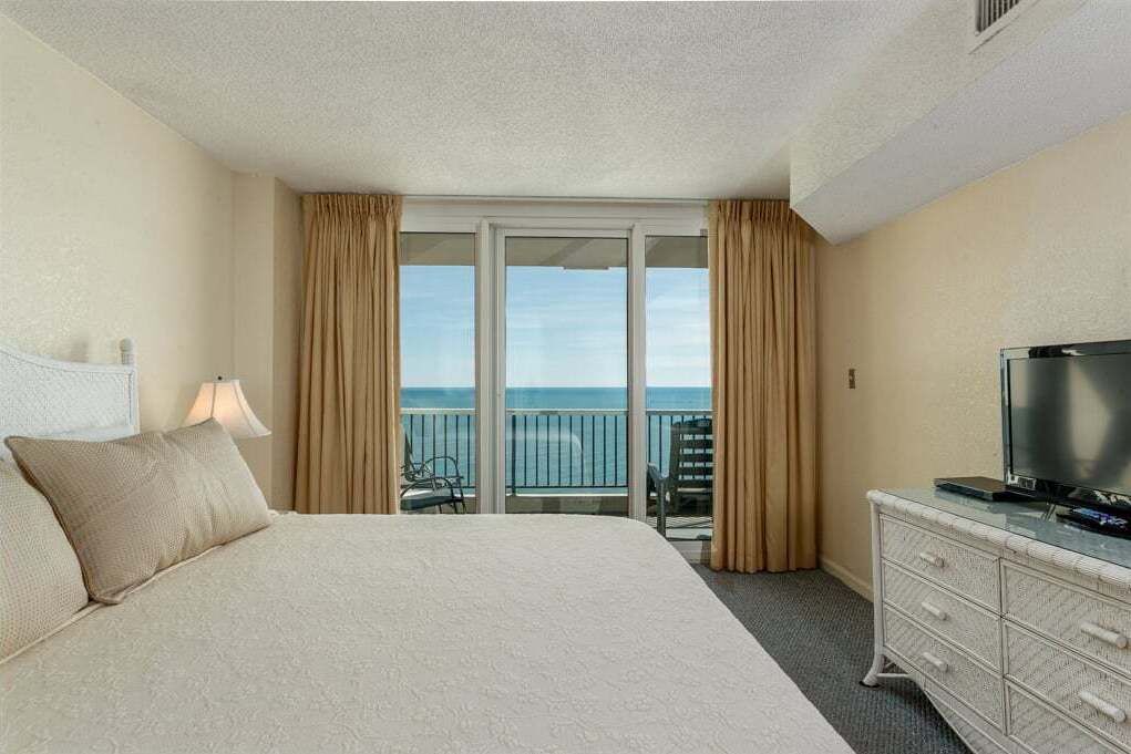 A hotel room with a large bed, a TV on a dresser, and a balcony with a view of the ocean.