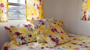 Cama com roupa de cama e almofadas florais, janela com cortinas florais e vista para árvores lá fora.