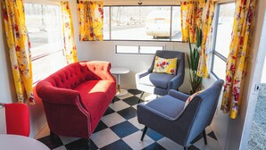 Um quarto com um sofá vermelho, uma poltrona cinza, uma mesa pequena branca e vista para uma motorhome branca pela janela.