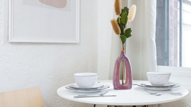 Ein runder Esstisch mit weißen Tellern und Schüsseln, eine rosa Vase mit Trockenpflanzen und ein gerahmtes Kunstwerk an der Wand.