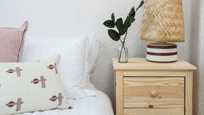 Ein ordentlich bezogenes Bett mit weißer Decke und Kissen, eine Nachttischlampe, ein kleiner Vasen mit Pflanze und ein gerahmtes Bild an der Wand.