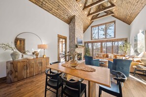 A spacious living area with a wooden dining table, black chairs, a stone fireplace, and a large window offering a view of trees. - Selah Vie at Eagles Nest - Vineyard Reserve-fire table, fire pit (Elk Park)