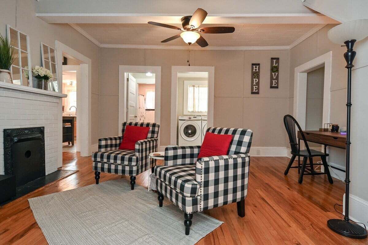 A living room with a fireplace, two checkered armchairs, a dining table, and a ceiling fan.