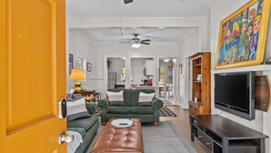 A living room with a sofa, ottoman, TV stand, and a painting. - Lenoir Cottage (Athens)