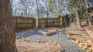 Un jardin avec une clôture en bois, un foyer extérieur, un espace salon aménagé et une zone recouverte de gravier et de pierres.