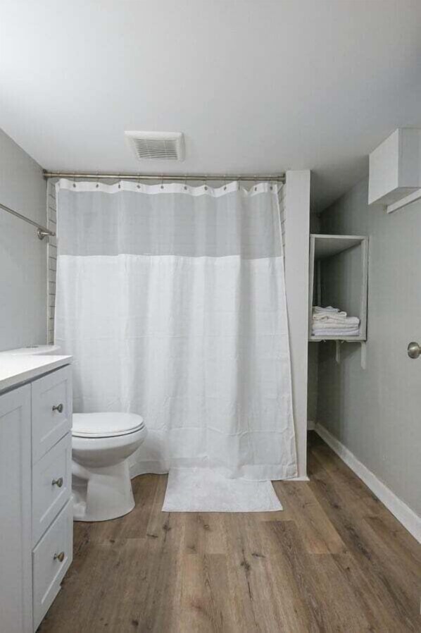 Une salle de bain avec des toilettes, un rideau de douche blanc, un plancher en bois et un meuble blanc contenant des serviettes.