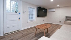 A room with a wooden floor, a white door, a window with blinds, a flat-screen TV mounted on the wall, and a round wooden table. - Bobbin Mills Studio - 1 Mile From Downtown Athens (Athens)