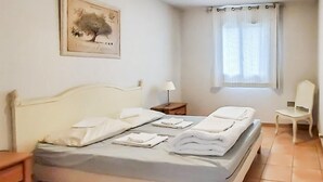 A neatly made bed with white linens and pillows, a bedside table with a lamp, a chair, and a window with curtains. - Amazing home in Montauroux with swimming pool (Montauroux)