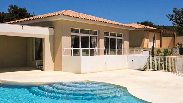 A swimming pool with a curved edge, a covered patio area, and a building with a tiled roof and large windows. - Amazing home in Montauroux with swimming pool (Montauroux)