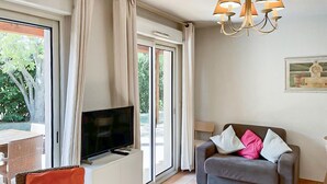 A living room with a TV on a white cabinet, a grey sofa with pillows, a chandelier, and a large window with curtains. - Amazing home in Montauroux with swimming pool (Montauroux)