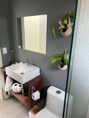 Un baño moderno con lavamanos, espejo y retrete blancos. Hay dos plantas en macetas en la pared y un estante de madera con artículos de tocador.