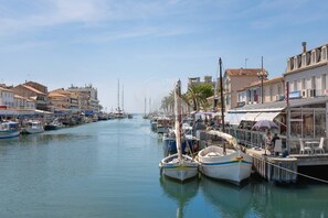A marina with boats docked along the water, buildings with shops and restaurants on the right, and a clear sky. - AS26823- P2 City Center - Side Sea View (LE GRAU DU ROI)