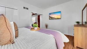 A modern bedroom with a bed, a wooden dresser, a painting, and a view into another room. - 3BR patio home w community pool, heated spa (Tucson)