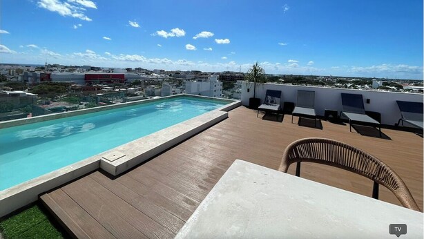 Una piscina en la azotea con vistas despejadas del paisaje urbano, una terraza de madera y sillones de descanso.