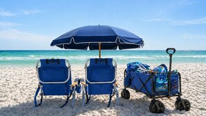 En strand med to blå strandstole og en parasol, en strandvogn, og kig til havet.