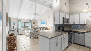 Una cocina amplia con armarios blancos, encimera de granito y una isla grande. Hay una zona de comedor con una mesa y sillas blancas, y una sala de estar con un sofá y una mesa de centro.