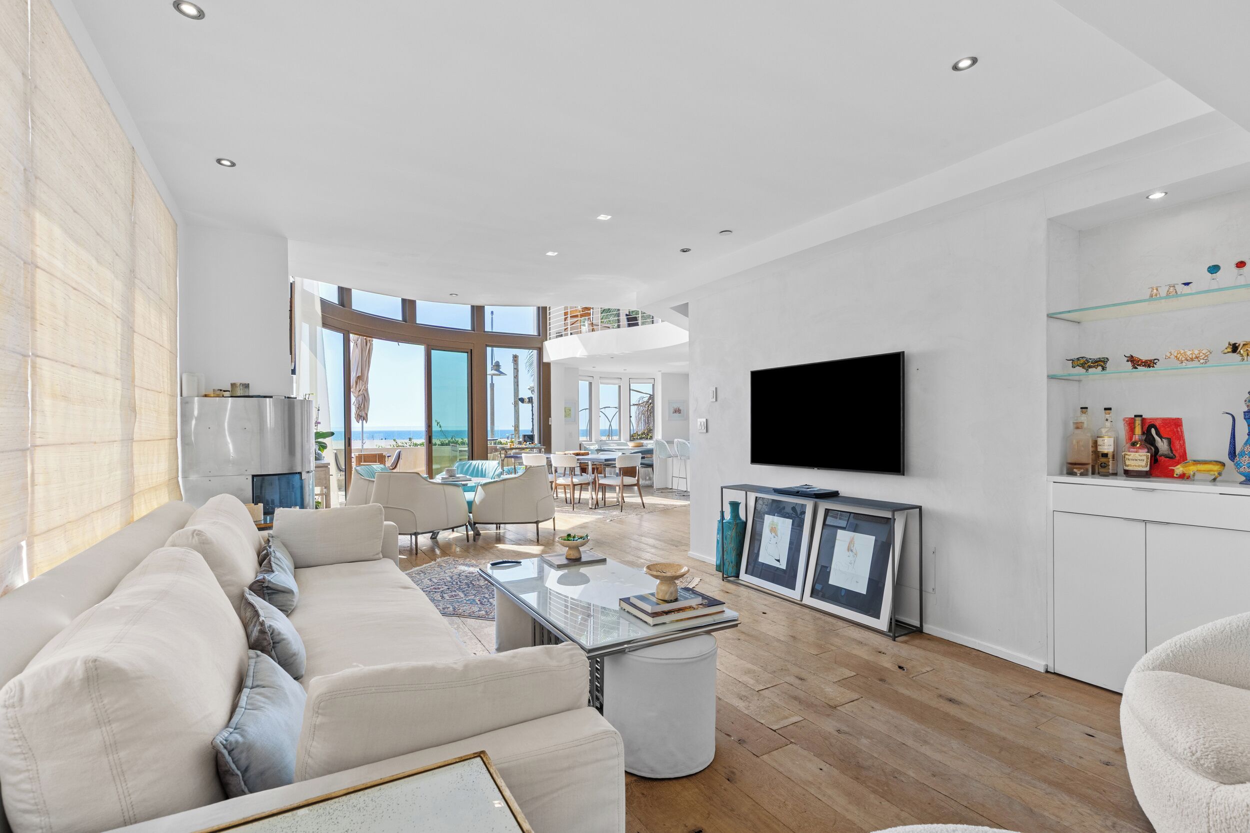 A spacious living room with a large sofa, a coffee table, and a flat-screen TV. There is a dining area with chairs and a table, and a kitchen area with a refrigerator and shelves.