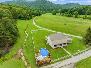 Una casa grande con patio cercado, piscina y vistas a un campo verde y colinas boscosas.