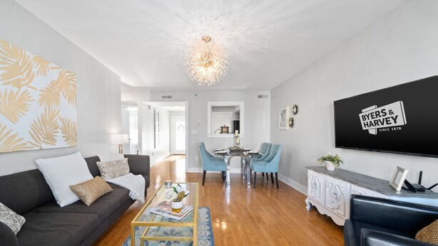 Una sala moderna con un sofá gris oscuro, una mesa de centro de vidrio y una zona de comedor con sillas azules. Un candelabro cuelga del techo, y un televisor de pantalla plana está montado en la pared.